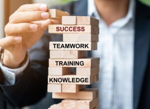 Hand removing a wooden block from a Jenga tower labeled SUCCESS, with other blocks reading TEAMWORK, TRAINING, and KNOWLEDGE beneath.